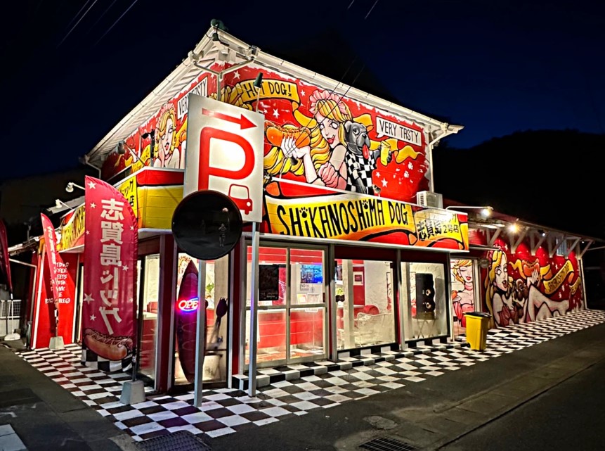 福岡市東区志賀島にある「志賀島ドッグ 2号店」の夜の外観。ライトアップされた赤い建物とアメリカンポップな壁画が映える店舗